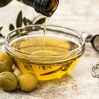 Close-up of olive oil being poured into a glass bowl surrounded by fresh olives and kitchen tools.