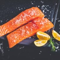 Close-up of raw salmon fillets garnished with salt, lemon wedges, and rosemary on a baking tray.